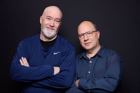 Two middle-aged men in navy shirts with arms crossed against a dark background, one bald with a grey beard and smiling, the other with glasses and a more serious expression